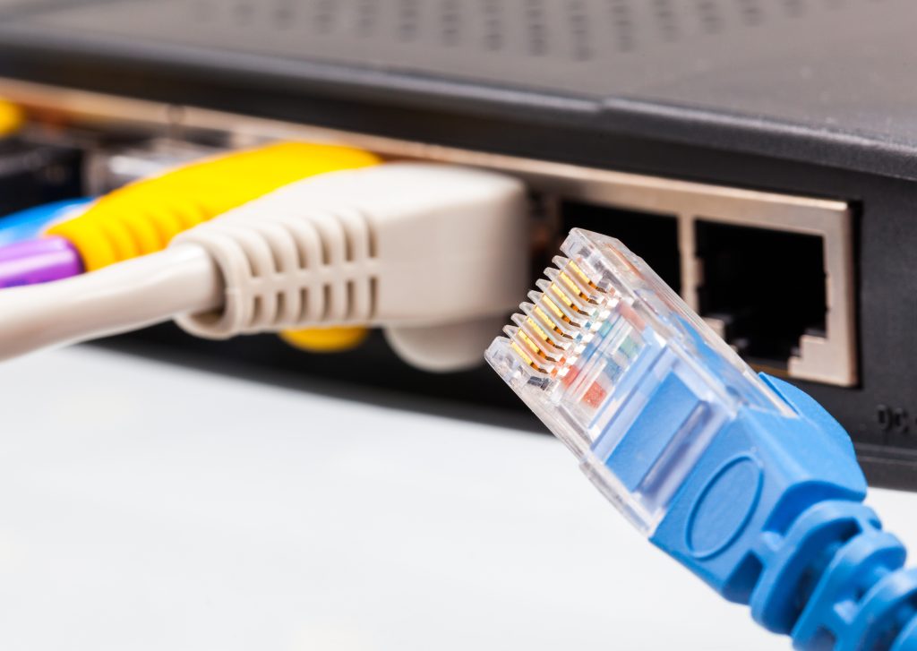 Close-up of a blue Ethernet cable with a visible connector, poised near an internet router with multiple colored cables plugged into its ports, ready to ensure seamless internet connectivity alongside the Wi-Fi network.