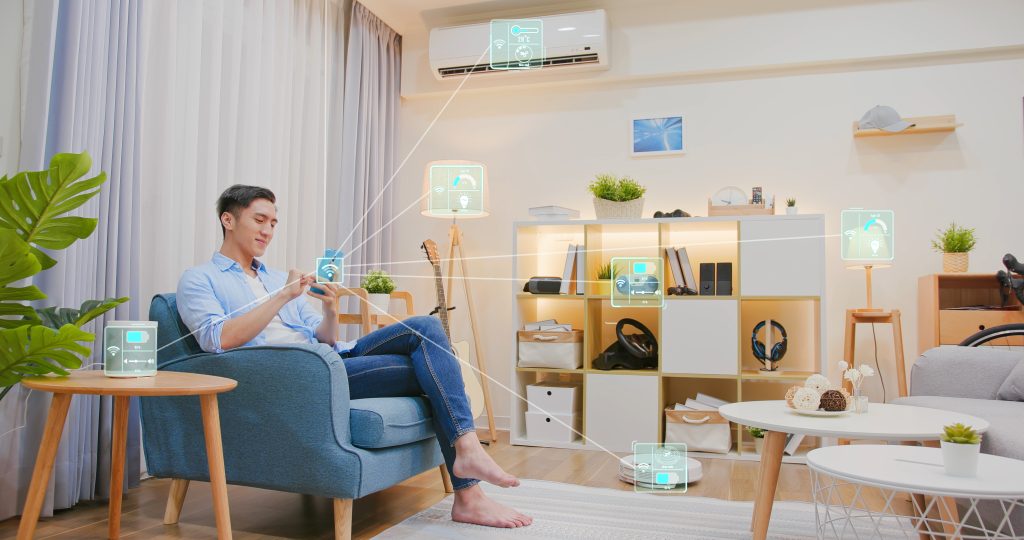 A person sits on a blue chair in a modern living room, using a smartphone to manage their smart home. Digital icons connected by lines represent devices like lights, air conditioner, and speakers. The bright room showcases a minimalist decor, perfectly blending technology and comfort.