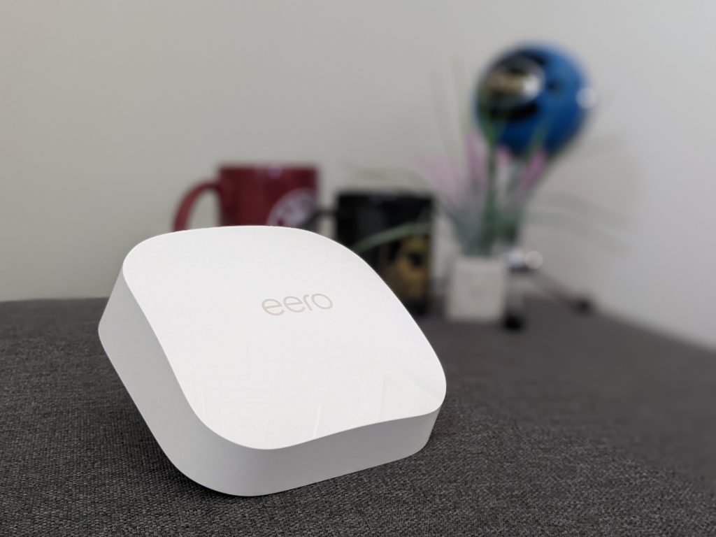 A white Eero mesh Wi-Fi router sits on a gray surface. In the blurred background, two mugs and a blue helmet-like object are visible.