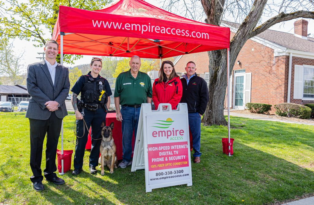 A group of five people and a police dog stand smiling under a red tent with "staging.empireaccess.com" on it, celebrating Empire Access's latest fiber expansion. A sign in front advertises their services. They are outdoors in front of a brick building.