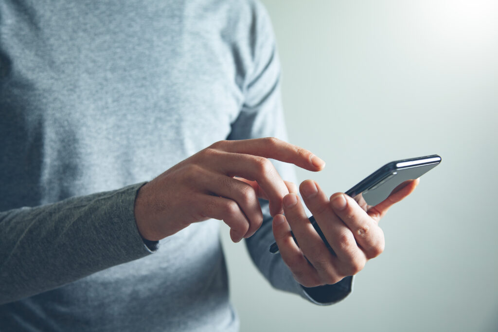 A person in a gray long-sleeve shirt is using a smartphone, tapping the screen to access their billing app. The background is plain and light-colored.