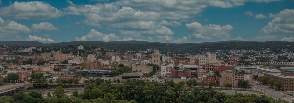 Aerial view of a cityscape with numerous brick buildings and the bustling energy of Scranton, surrounded by lush green hills under a partly cloudy blue sky.