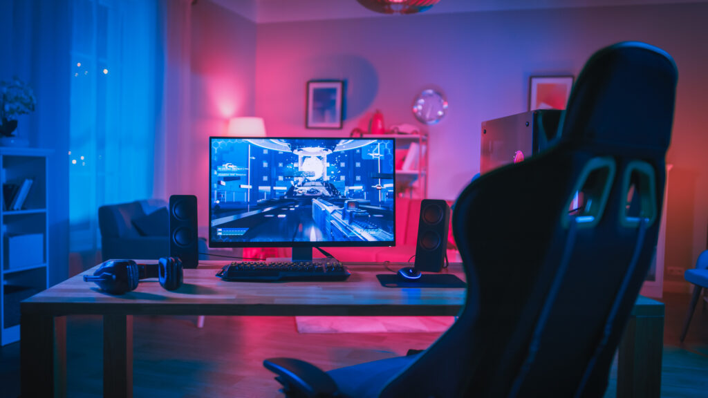 A gaming setup with a computer monitor displaying a futuristic game immerses you in an electrifying atmosphere. The room glows with blue and pink ambient lighting, while a gaming chair, keyboard, speakers, and headphones sit on the wooden desk. Cozy interior touches include a sofa and framed pictures in the background.