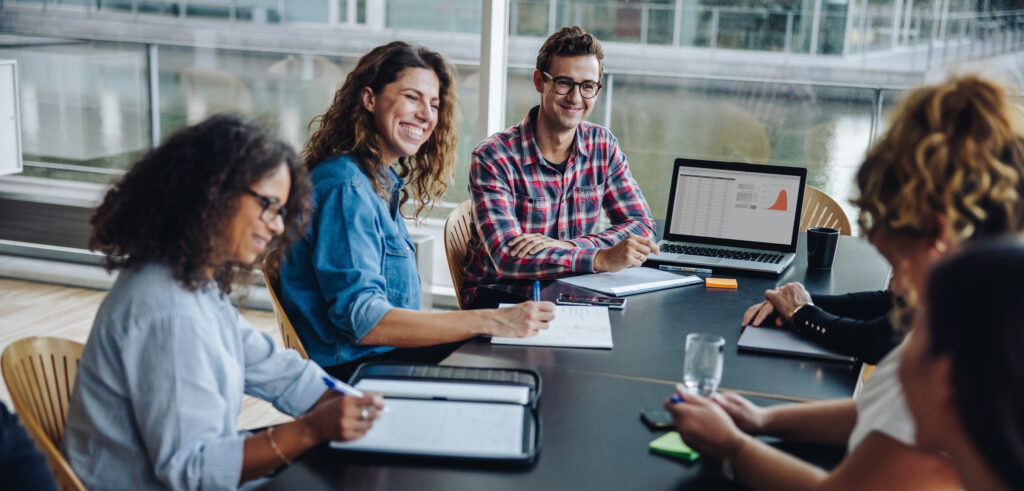 A woman and four colleagues sit around a conference table, smiling and engaged in discussion. Jane is using her laptop to display a pie chart. Notebooks and pens scattered on the table suggest a collaborative meeting or brainstorming session.