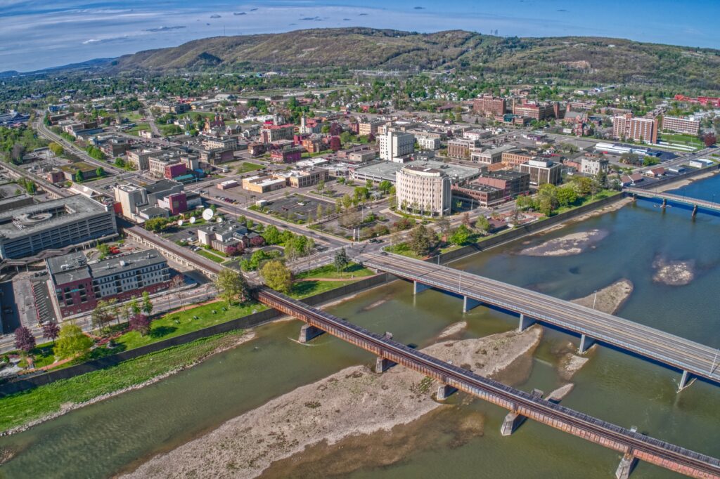 An aerial view of a city expands across several bridges that span a wide river. The cityscape, with its diverse buildings, roads, and lush green areas, unfolds against a backdrop of hills beneath a partly cloudy sky.