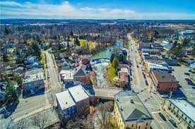 Aerial view of Bloomfield, a charming small town with streets lined by various buildings. A river meanders through, embraced by lush trees and greenery. The sky is clear with a few clouds, and the horizon boasts distant hills and fields.