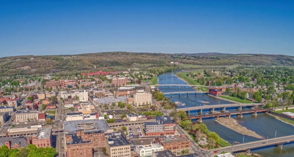 An aerial view of a town showcases several bridges crossing a river as it expands through the area. The scene features buildings, roads, and a mix of greenery, with rolling hills visible in the background under a clear blue sky.