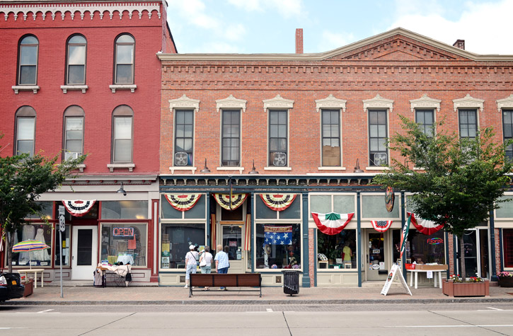 A historic street with red brick buildings features American flags and patriotic bunting. People stroll along the sidewalk, enjoying the various services offered by quaint shops. A bench invites visitors to relax as trees line the way on a clear day.
