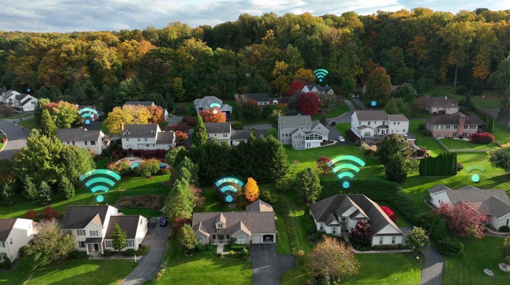 An aerial view of a suburban neighborhood shows several houses surrounded by trees. Symbols depicting wireless signals hover above rooftops, indicating internet connectivity. Local businesses are nestled among the homes, with autumn foliage providing a vibrant backdrop.