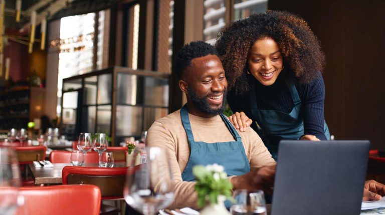 A man and woman, both wearing aprons, stand behind a laptop in a cozy restaurant setting. They are smiling and appear engaged with the screen. The table is set with glasses and a small plant centerpiece.