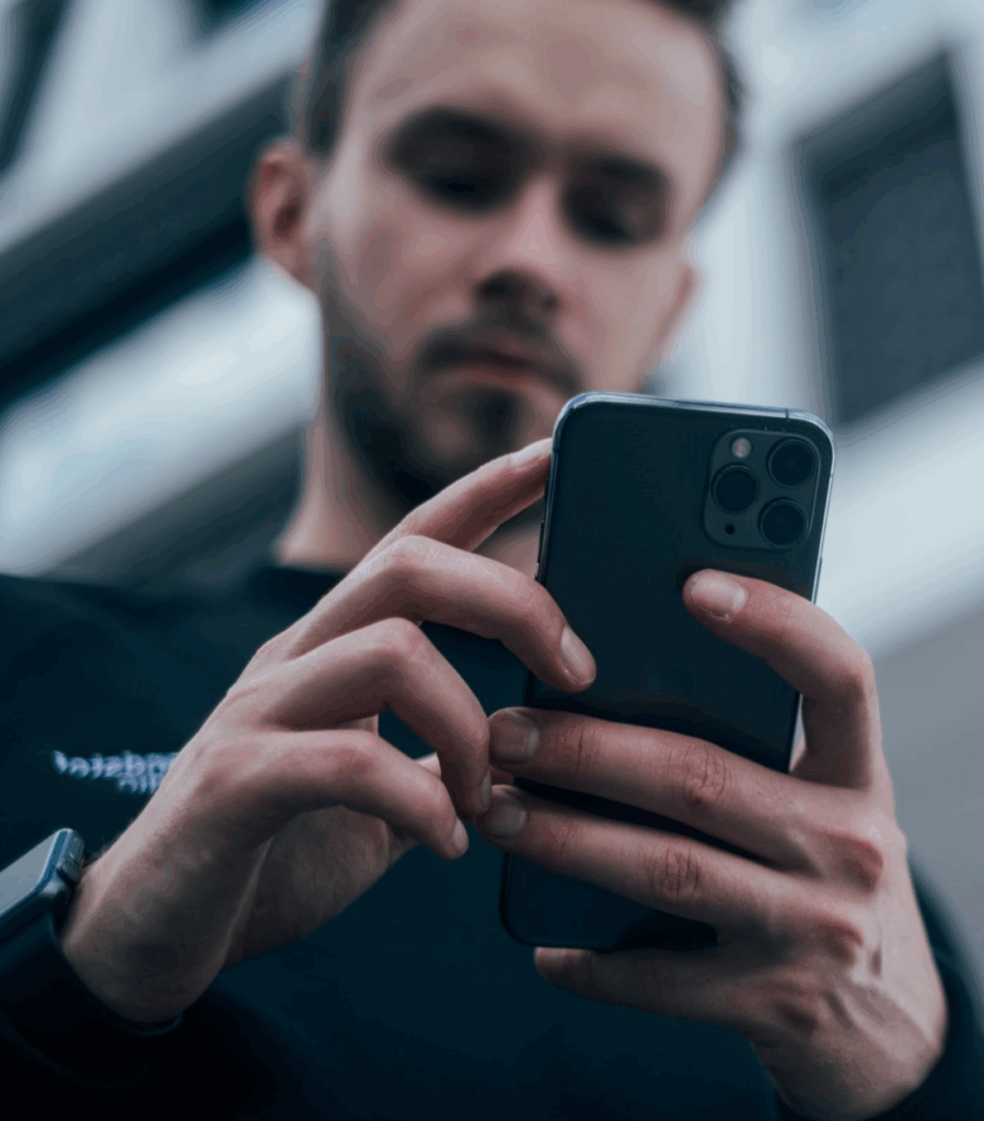 A person wearing a dark shirt and smartwatch is holding and using a smartphone, with the photo focused on their hands and phone, and their face slightly blurred in the background.